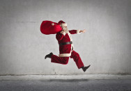 Ein Mann verkleidet als Weihnachtsmann springt in der Luft und trägt einen großen roten Sack auf der Schulter. Der Hintergrund ist eine einfache graue Wand.