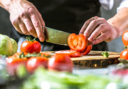 Ein Mann schneidet eine Tomate mit einem scharfen Küchenmesser auf einem Holzbrett. Im Vordergrund und Hintergrund sind weitere Tomaten und grünes Gemüse zu sehen.