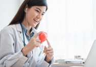 Eine Ärztin sitzt an einem Schreibtisch und trägt Kopfhörer, während sie mit einem Patienten über einen Laptop spricht. Sie hält ein rotes Modell eines Herzens in der Hand und erklärt möglicherweise eine medizinische Angelegenheit.