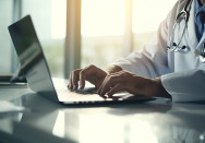 Ein Arzt in einem weißen Kittel mit Stethoskop um den Hals arbeitet konzentriert an einem Laptop in einer modernen, hellen Arztpraxis.