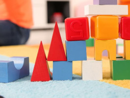 Bauklötze bilden einen Turm in einem Spielzimmer. Im Hintergrund sitzt ein Kind im Schneidersitz und schaut nach links.