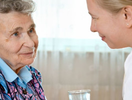Pflegekraft spricht liebevoll mit einer älteren Frau im Rollstuhl und reicht ihr ein Glas Wasser.