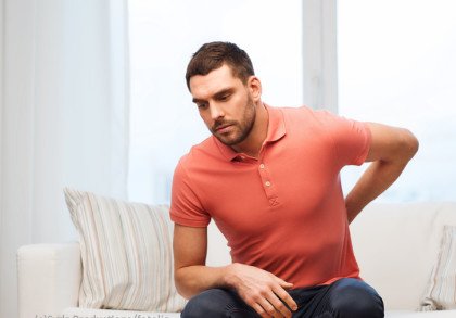 Ein Mann trägt ein lachsfarbenes Polohemd und sitzt auf einem weißem Sofa. Er greift sich an den unteren Rücken und zeigt ein schmerzverzerrtes Gesichtsausdruck, was auf Rückenschmerzen hindeutet.