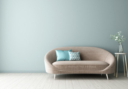 Ein modernes Wohnzimmer mit einer taupefarbenen Couch, die sich vor einer mintgrün gestrichenen Wand befindet. Zwei Dekokissen in Türkis und mit geometrischem Muster zieren die Couch.
