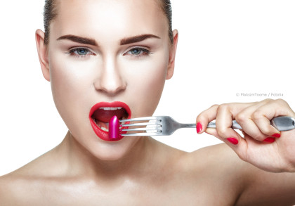 Eine Frau hält eine Gabel mit einem Stück pinkem Lippenstift vor ihren geöffneten Mund. Sie trägt roten Lippenstift und hat lackierte Nägel in der gleichen Farbe. Der Hintergrund ist weiß.