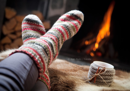 Ein Paar Füße in bunt gestreiften Wollsocken vor einem Kaminfeuer. Im Vordergrund liegt eine Tasse Tee oder Kaffee mit einem gestrickten Überzug auf einem Fell. Im Hintergrund sind Holzscheite zu sehen.