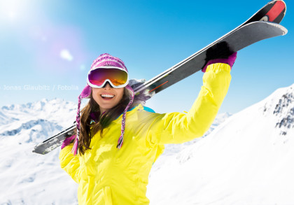 Frau in bunter Winterkleidung trägt Ski auf ihrer Schulter und lächelt in die Kamera. Sie trägt eine pink gestreifte Mütze, eine gelbe Skijacke, Skibrille und Handschuhe. Im Hintergrund sind schneebedeckte Berge und ein klarer, blauer Himmel zu sehen.