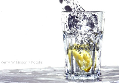 Ein Glas Wasser mit einer Zitronenscheibe, in das Wasser gegossen wird. Das Wasser spritzt und erzeugt Bewegung im Glas.