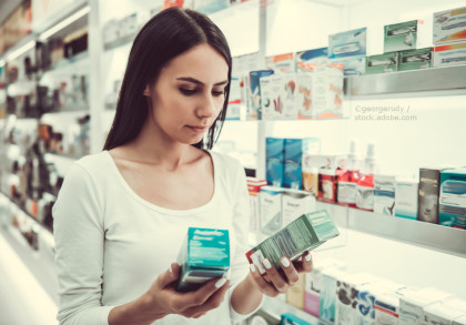 Eine junge Frau steht in einer Apotheke und hält zwei Medikamentenschachteln in den Händen, während sie diese aufmerksam betrachtet. Im Hintergrund sind zahlreiche weitere Medikamentenschachteln auf Regalen zu sehen.