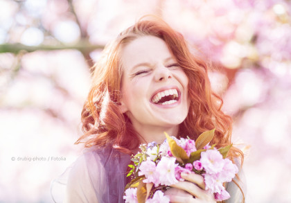 Eine rothaarige Frau lacht herzhaft und hält einen Strauß pinker Blumen in den Händen. Im Hintergrund sind verschwommene, blühende Bäume zu sehen.
