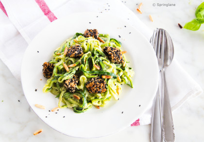 Ein weißer Teller mit Zucchini-Nudeln, garniert mit gerösteten Pinienkernen und knusprigen Stücken in schwarzem und weißem Sesam. Daneben liegen eine Gabel und ein Löffel auf einer weißen Serviette mit roten Streifen.
