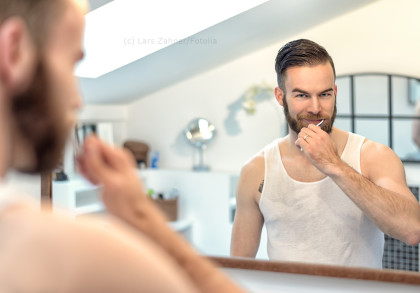 Ein Mann mit Bart steht vor einem Spiegel und putzt seine Zähne. Er trägt ein weißes Tanktop und schaut lächelnd in den Spiegel. Der Raum ist hell, mit modernen Badezimmeraccessoires im Hintergrund.