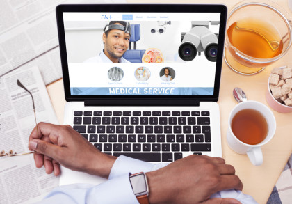 Ein Laptop zeigt eine Medizin-Website mit einem lächelnden Arzt auf dem Bildschirm. Eine Person hält eine Brille in der linken Hand und gibt mit der rechten Hand die Tastatur ein.