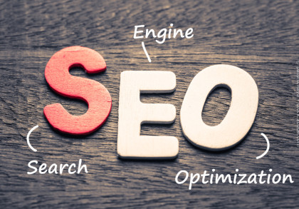 Holzbuchstaben auf einem dunklen Holztisch, die das Wort "SEO" bilden. Das "S" ist rot, während das "E" und das "O" weiß sind. Um die Buchstaben stehen die Wörter "Search", "Engine" und "Optimization" in weißer Schrift, die das Akronym SEO erklären.