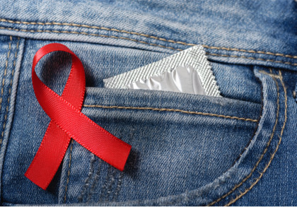 Ein rotes Band, das als Symbol für die Unterstützung der AIDS-Bekämpfung dient, ist auf eine Jeans-Tasche gesteckt. In der Tasche steckt ein silbernes Kondompäckchen.