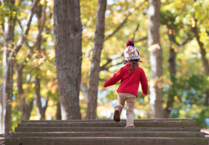 Ein kleines Kind in rotem Pullover und bunter Wintermütze läuft eine Treppe hinauf in einem herbstlichen Wald mit bunten Blättern an den Bäumen.