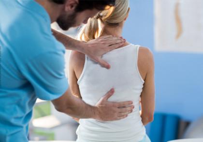 Ein Physiotherapeut in einem blauen Hemd massiert den Rücken einer Frau mit blonden Haaren, die ein weißes ärmelloses Top trägt. Die Frau sitzt und der Physiotherapeut steht hinter ihr, während er ihre Schultern und den unteren Rückenbereich bearbeitet.