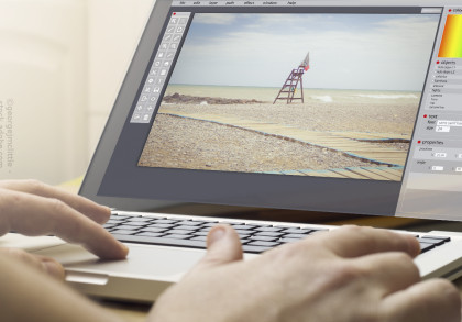 Eine Person arbeitet an einem Laptop, auf dem ein Bildbearbeitungsprogramm geöffnet ist. Auf dem Bildschirm ist ein bearbeitetes Foto von einem leeren Strand mit einem Rettungsschwimmerturm zu sehen. Der Strand hat Kieselsteine und Holzstegwege.