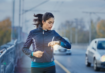 Eine Frau joggt auf einer Straße und schaut auf ihre Armbanduhr. Sie trägt eine schwarze Jacke und ein blaues Sportoberteil. Im Hintergrund sind Autos und leicht verschwommene Bäume zu sehen.