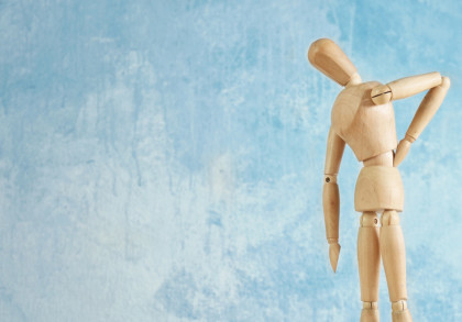 Eine Holzfigur, die Rückenschmerzen darzustellen scheint, steht vor einem blauen Hintergrund. Die Figur hat eine Hand auf den Rücken gelegt und beugt sich leicht nach vorne.