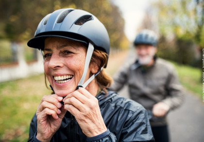 Eine ältere Frau mit einem grauen Fahrradhelm lacht und befestigt den Riemen ihres Helms. Im Hintergrund ist ein älterer Mann, ebenfalls mit Helm, unscharf zu sehen. Die beiden befinden sich auf einem Weg im Freien, umgeben von grüner Natur.