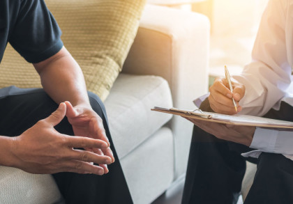 Das Bild zeigt einen Patienten und einen Arzt in einer Beratungssituation. Der Patient sitzt auf einer Couch und hält seine Hände verschränkt vor seinem Körper. Der Arzt sitzt mit einem Klemmbrett und einem Stift in der Hand, bereit, Notizen zu machen.