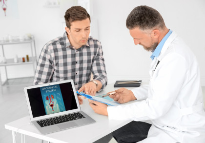 Ein Arzt im weißen Kittel bespricht mit einem Patienten in einem karierten Hemd einen Bericht. Auf dem Tisch vor ihnen befindet sich ein Laptop, auf dessen Bildschirm eine Abbildung des Urogenitalsystems zu sehen ist.