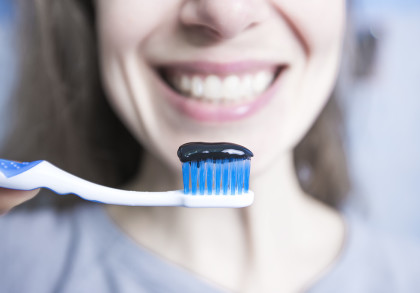 Nahaufnahme eines lächelnden Gesichts, das eine Zahnbürste mit blauen Borsten und schwarzer Zahnpasta darauf hält.