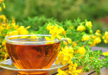 Eine Tasse Kräutertee neben frischen gelben Johanniskrautblüten auf einem Holztisch im Freien. Im Hintergrund sind weitere grüne Pflanzen und verschwommene gelbe Blüten zu sehen.