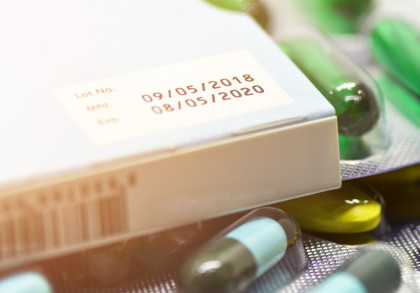 Nahaufnahme einer Medikamentenschachtel mit dem Herstellungsdatum "09/05/2018" und dem Verfallsdatum "08/05/2020". Umliegend sind Blisterpackungen mit grünen, gelben und blau-grünen Kapseln und Tabletten zu sehen.