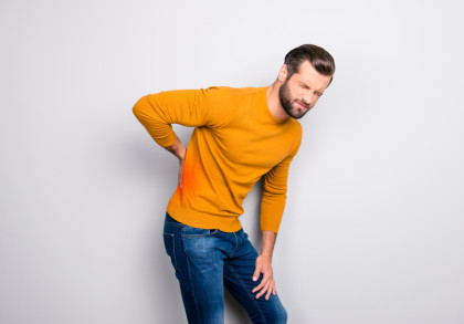 Ein Mann im gelben Pullover und blauer Jeans hat Schmerzen im unteren Rückenbereich. Er hält seine Hand auf die schmerzende Stelle und verzieht das Gesicht vor Schmerzen vor einem weißen Hintergrund.