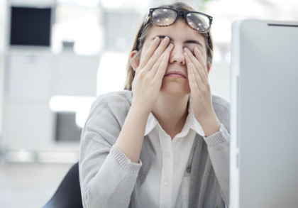 Eine Frau sitzt vor einem Computerbildschirm und reibt sich müde die Augen. Sie trägt ein weißes Hemd und eine graue Strickjacke. Eine Sonnenbrille ist auf ihren Kopf geschoben. Der Hintergrund ist unscharf und zeigt ein Büro oder einen Arbeitsbereich.