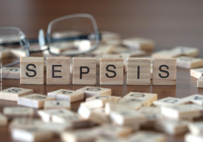 Auf dem Bild sind Holzbuchstabenblöcke zu sehen, die das Wort "SEPSIS" bilden. Die Blöcke liegen auf einem Holztisch. Im Hintergrund sind andere Buchstabensteine verstreut und eine Brille ist leicht unscharf zu erkennen.