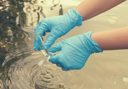 Nahaufnahme von zwei Händen mit blauen Einweghandschuhen, die Wasserproben aus einem Teich oder See entnehmen.