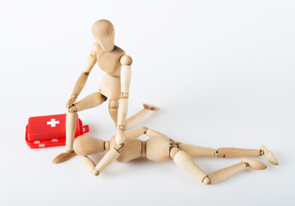 Holzpuppen-Figuren, die eine Erste-Hilfe-Situation darstellen. Eine Figur kniet neben einer anderen, die auf dem Boden liegt, und streckt eine Hand aus. Neben ihnen liegt ein roter Erste-Hilfe-Kasten mit einem weißen Kreuz. Der Hintergrund ist weiß.