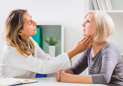 Eine Ärztin untersucht den Hals einer älteren Patientin in einem hellen, modernen Behandlungsraum. Die Ärztin trägt einen weißen Kittel und die Patientin ein graues Oberteil.