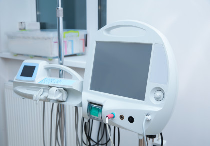 Medizinisches Gerät in einem Krankenhauszimmer. Links ein kleineres Steuergerät mit Display und Tasten, rechts ein größeres mit großem Bildschirm. Kabel und Schläuche sind angeschlossen, im Hintergrund ist ein Fenster und eine Box mit Utensilien.
