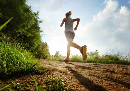 Eine Person joggt auf einem sandigen Pfad im Freien. Die Sonne scheint durch die Bäume und Pflanzen auf der linken Seite, während am Himmel einige Wolken zu sehen sind. Die Person trägt sportliche Kleidung und Laufschuhe.