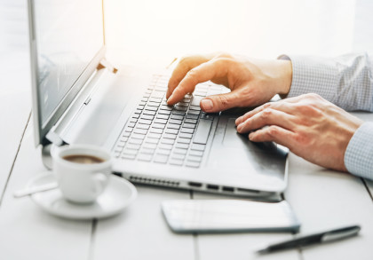 Nahaufnahme von Händen, die auf einem Laptop-Tastatur tippen. Auf dem Tisch stehen eine weiße Kaffeetasse mit Untertasse und Löffel, ein Stift und ein Smartphone. Sanftes Sonnenlicht fällt von oben links auf die Szene. Eine helle, produktive Atmosphäre.