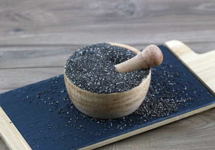 Ein Holzmörser und -stößel gefüllt mit Chiasamen steht auf einem schwarzen Schieferbrett, das auf einem Holzschneidebrett liegt. Einige Chiasamen sind um den Mörser herum verstreut. Der Hintergrund besteht aus einer holzartigen Oberfläche.