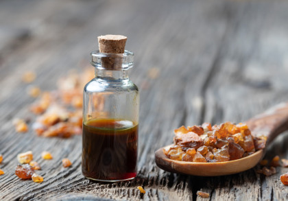 Eine kleine Glasflasche mit einem Korkverschluss, die eine dunkle Flüssigkeit enthält, steht auf einem holzernen Hintergrund. Daneben liegen auf einem hölzernen Löffel einige kristalline Stücke, wahrscheinlich Harz- oder Bernsteinfragmente.