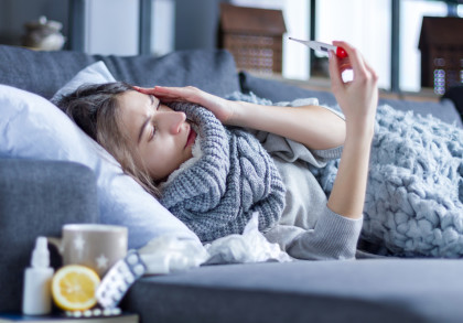 Eine Frau liegt krank auf einem Sofa, eingehüllt in eine graue Decke und einen Schal. Sie hält sich den Kopf und schaut auf ein Thermometer in ihrer Hand.