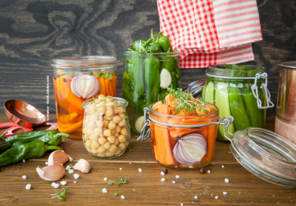 Glasgefäße mit eingelegtem Gemüse auf einem Holztisch; enthaltene Zutaten sind marinierte Karotten, Kichererbsen, grüne Paprika und Gurken. Einige Knoblauchzehen, grobkörniges Salz und eine rot-weiß karierte Stoffserviette sind ebenfalls zu sehen.
