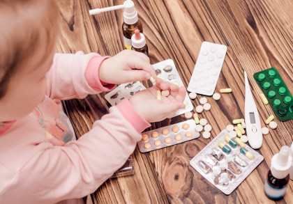 Kleines Kind im rosa Schlafanzug sitzt an einem Holztisch & hält eine gelbe Kapsel in der Hand. Auf dem Tisch liegen verschiedene Medikamente, darunter Tabletten in Blisterpackungen, Kapseln, ein digitales Fieberthermometer & medizinische Tropfflaschen.
