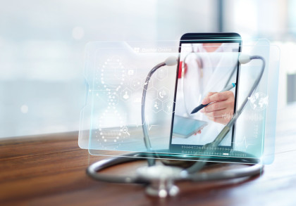 Ein Stethoskop liegt auf einem Holztisch, während im Hintergrund ein Tablet oder Smartphone zu sehen ist, das einen Arzt zeigt, der auf einem digitalen Gerät notiert.