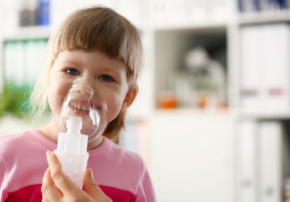 Ein kleines Mädchen in einem rosa Oberteil verwendet einen Inhalator mit einer Gesichtsmaske. Sie lächelt in die Kamera und befindet sich in einem gut beleuchteten Raum mit Regalen im Hintergrund.