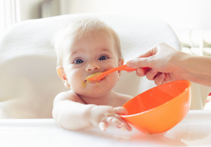 Ein Baby sitzt in einem Hochstuhl und wird mit einem orangefarbenen Löffel aus einer orangefarbenen Schüssel gefüttert. Das Baby hat ein wenig Essen um den Mund herum. Eine Hand hält den Löffel, während das Baby zuschaut und die Schüssel festhält.