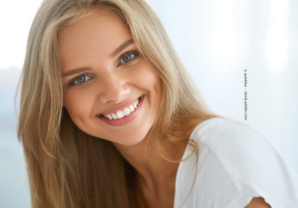 Eine junge Frau mit langen blonden Haaren und blauen Augen lächelt in die Kamera. Sie trägt ein weißes Oberteil. Der Hintergrund ist verschwommen und in hellen Farben gehalten.