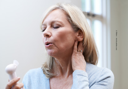 Eine ältere Frau hält sich den Nacken und benutzt einen kleinen Handventilator, um sich abzukühlen. Sie trägt einen hellblauen Pullover und hat blonde Haare. Sie steht in einem hellen Raum.