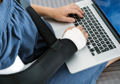 Eine Person mit einem blauen Oberteil tippt auf einer Laptop-Tastatur. Ihr rechter Arm ist in einem Verband und einer Armschlinge fixiert.